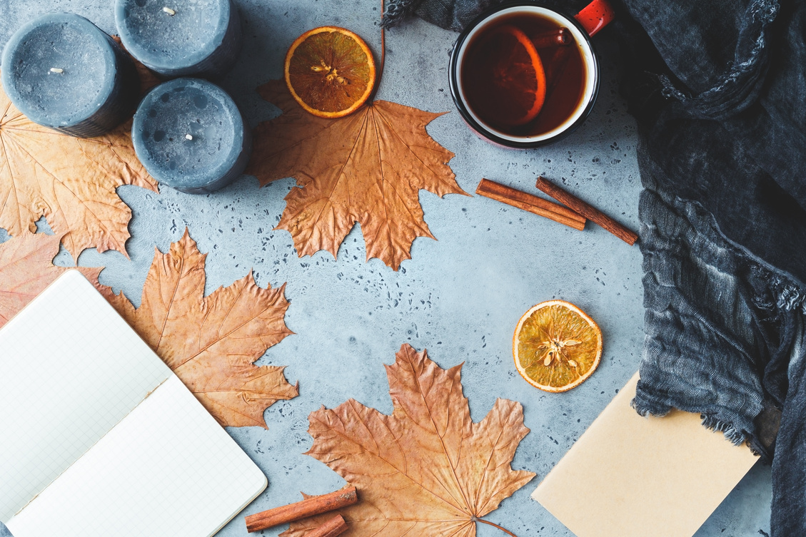 Autumn frame flat lay composition on a grey concrete background. Maple leaves, season tea,   open notebook with empty sheets, black aromatic candles and warm scarf.